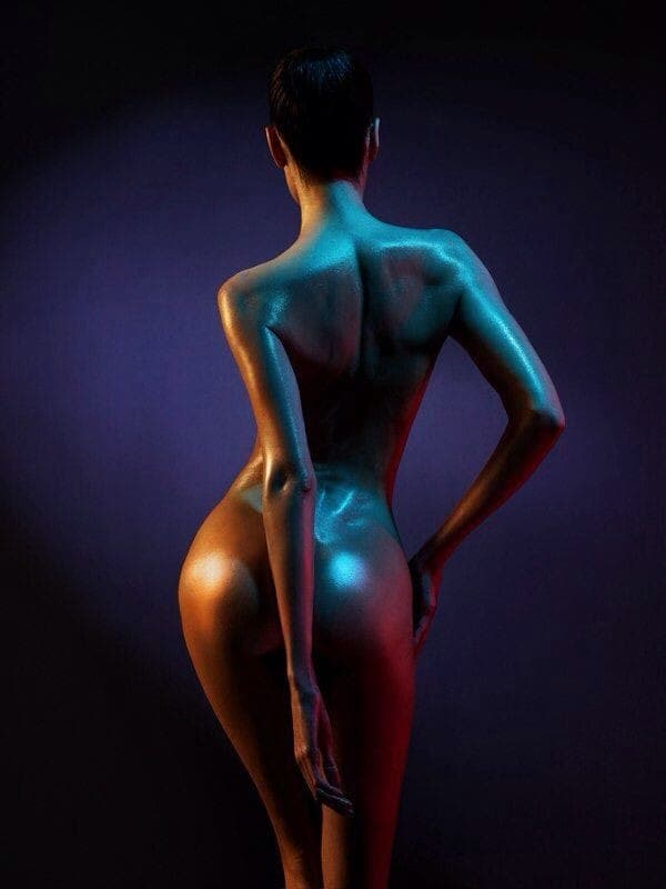 A person with short hair stands with their back to the camera, illuminated by dramatic blue and red lighting, evoking the serene atmosphere of a tantric massage and highlighting the contours and muscle definition of their nude body.