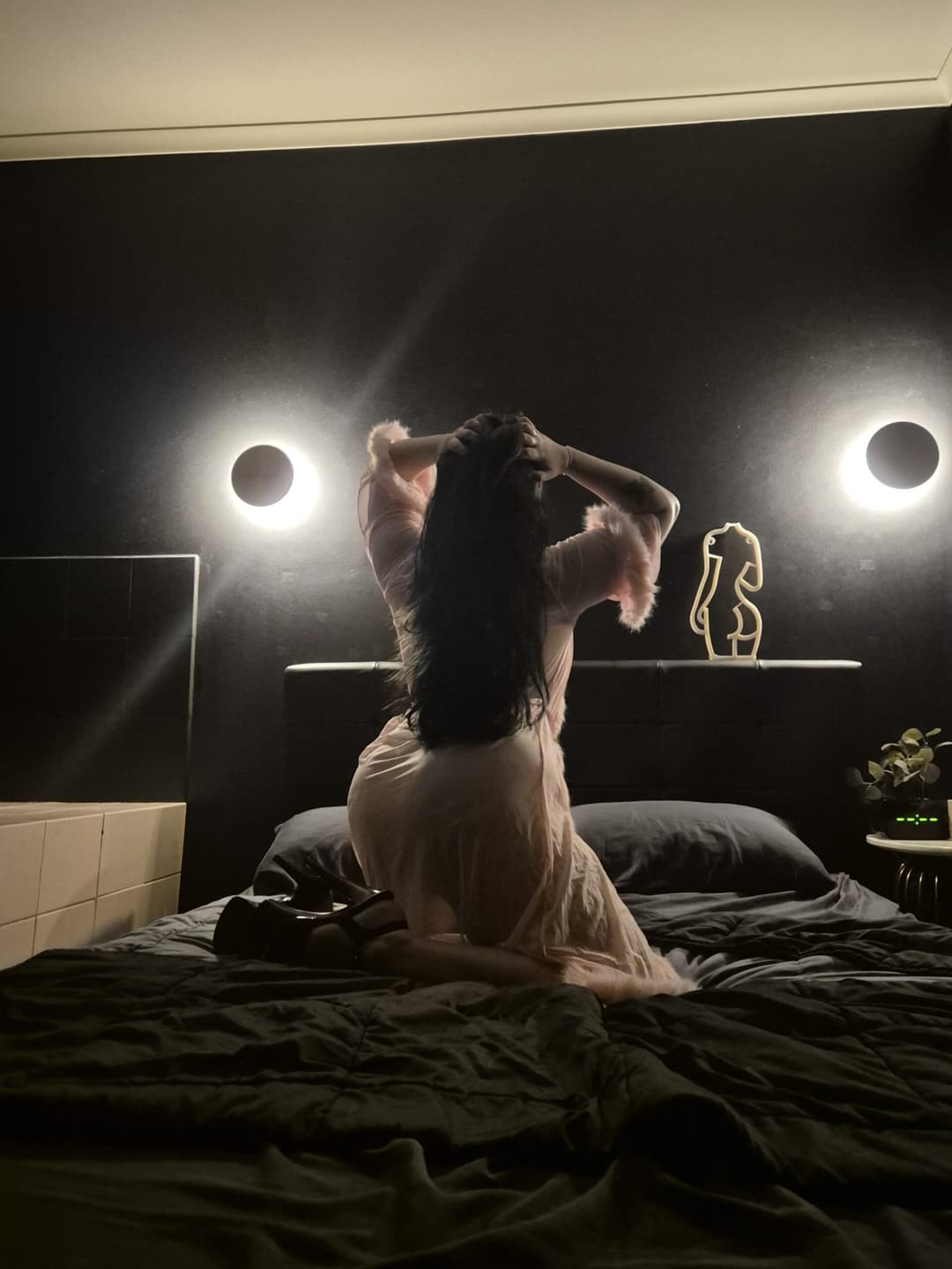 A person in a light pink dressing gown kneels on a bed, facing a dark wall with two round lights. The room has a moody, dim atmosphere—perfect for an independent masseuse offering a relaxing sensual massage experience.