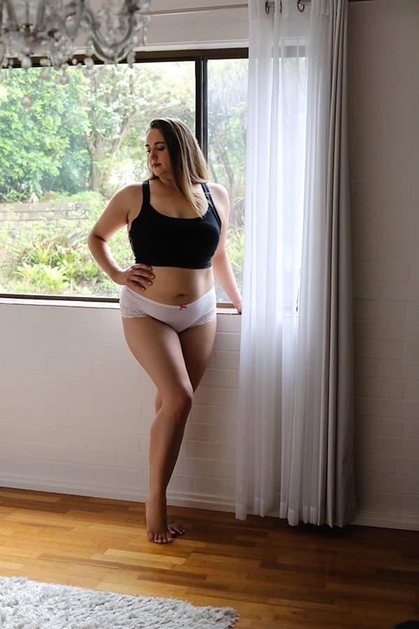 A woman in a black bra and white knickers stands barefoot by a window with sheer white curtains, looking outside. The tranquil scene evokes the calming ambience often found with an independent masseuse. Wooden floor and greenery visible through the window.