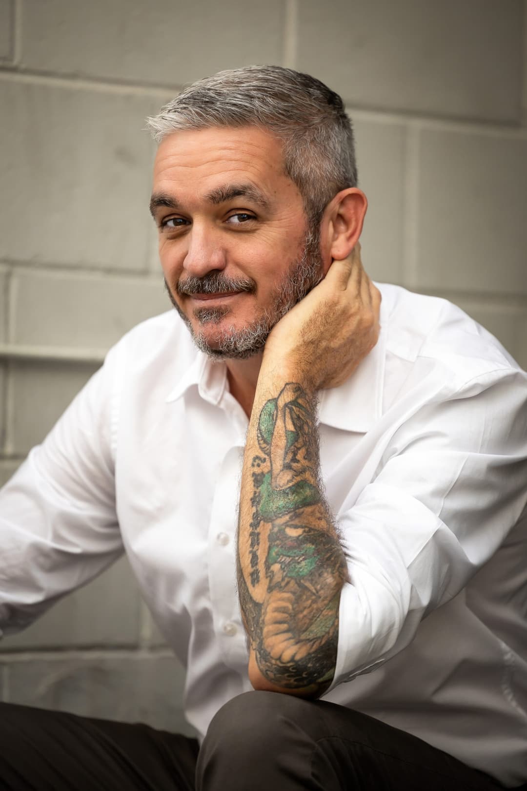 A man with short grey hair and a beard, wearing a white shirt, sits in front of a light brick wall. Smiling warmly and resting his head on his hand, he reveals a colourful tattoo—exuding the relaxed vibe of a tantric massage Australia expert.