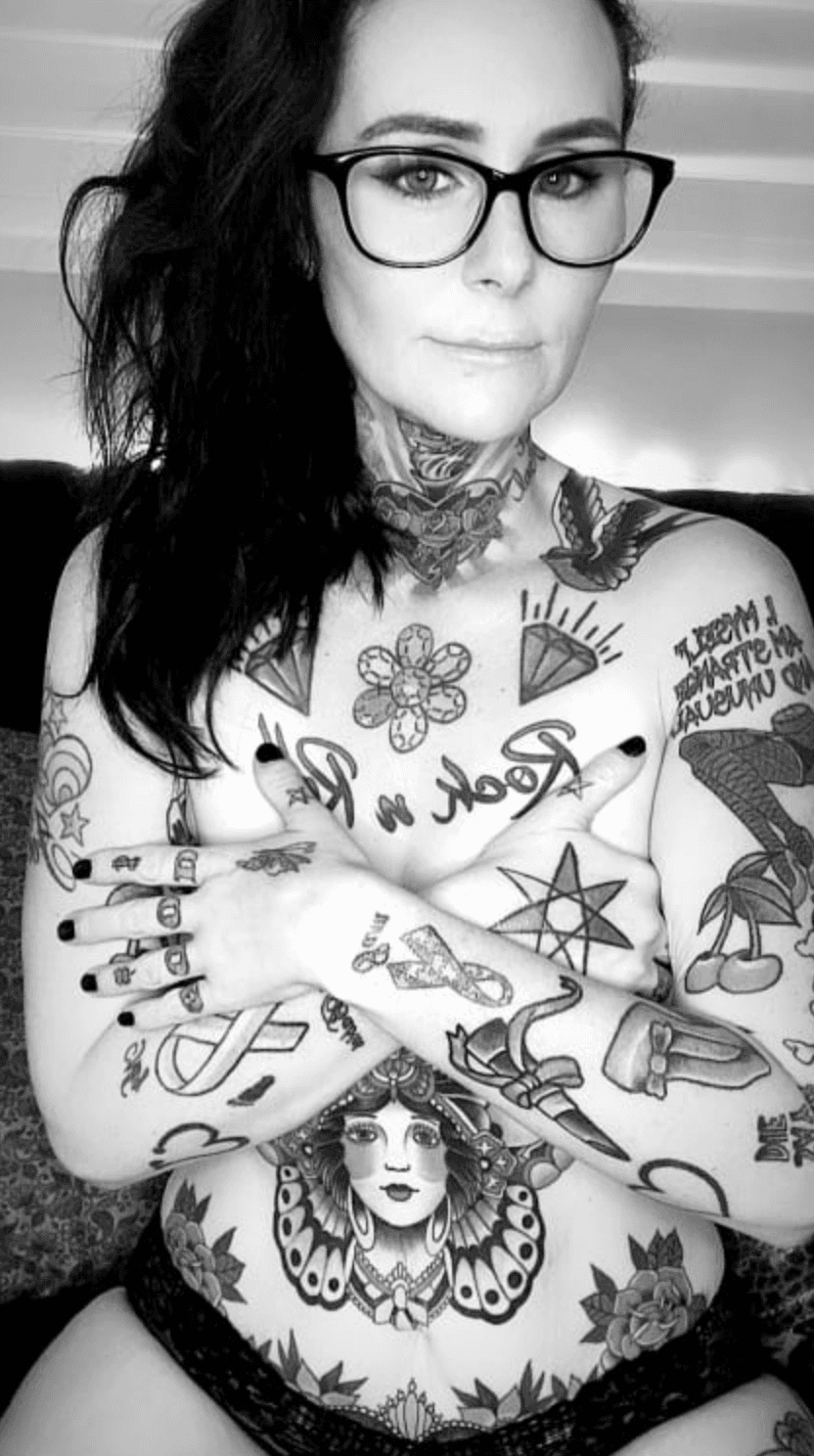 A woman with long dark hair and glasses poses confidently with her arms folded, displaying numerous tattoos on her arms, torso, and neck. She is wearing dark underwear in this black and white photo evoking the edgy allure of a milking table massage setting.