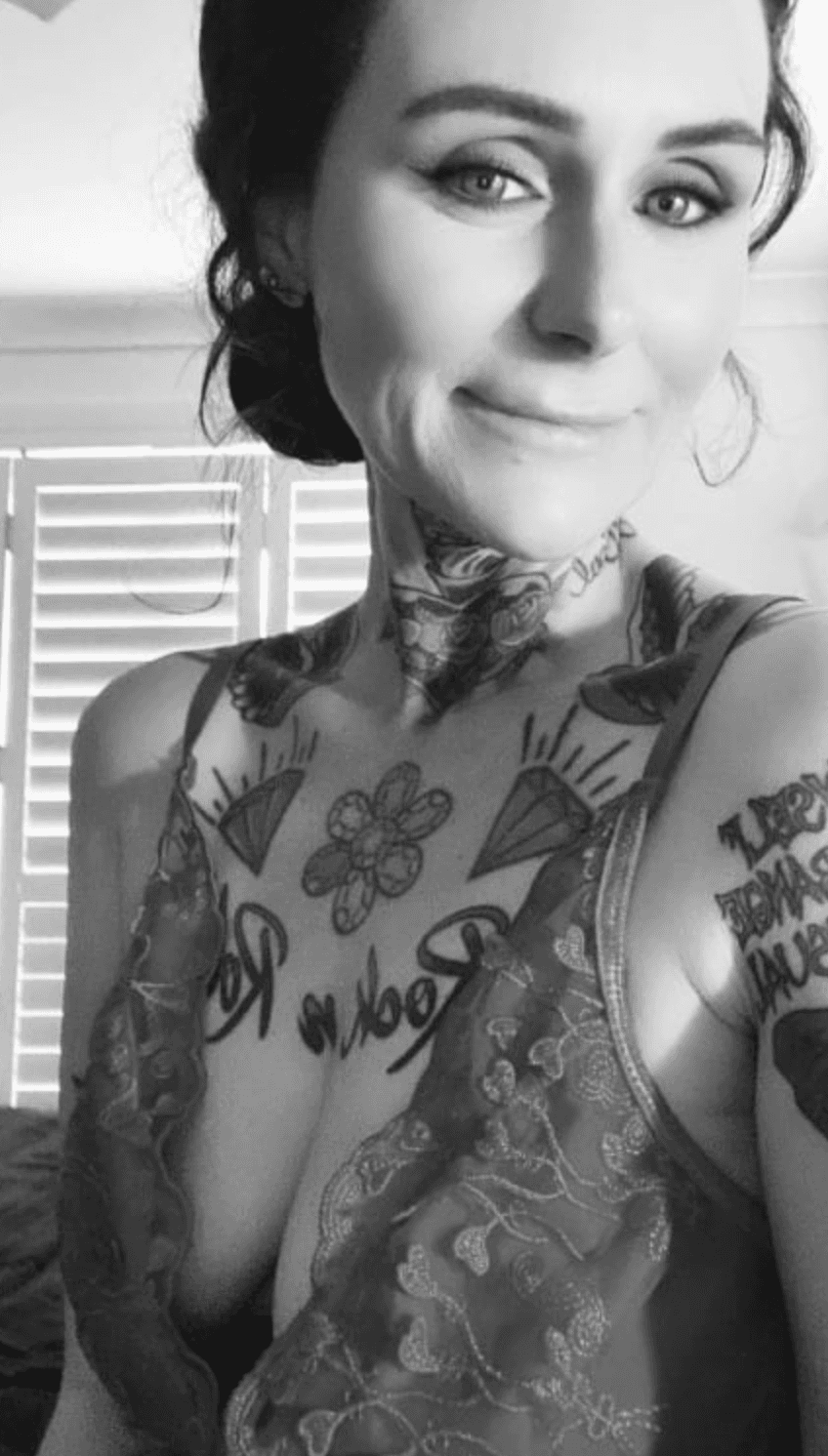 Black and white photo of a woman with dark hair, smiling slightly. She wears a lace top and shows multiple tattoos on her chest, neck, and arm. Blinds and a window are visible in the background, evoking the calm of a milking table massage space.