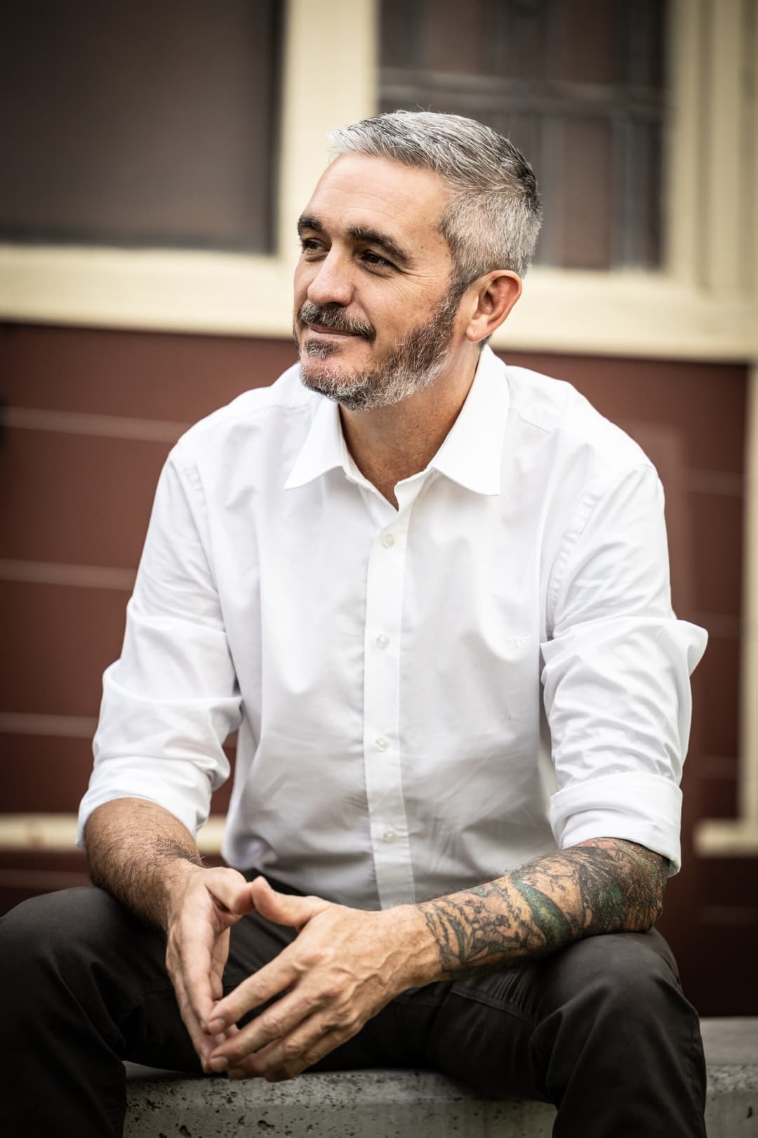 A man with short grey hair and a beard, wearing a white button-up shirt and dark trousers, sits outdoors, smiling thoughtfully. As an independent masseur specialising in sensual massage, he displays a tattoo sleeve on his left forearm.