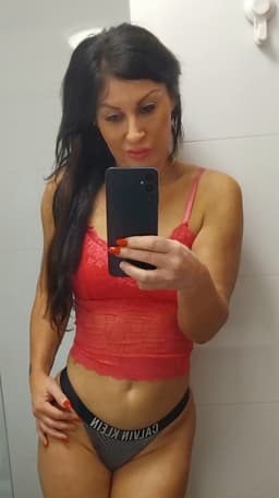 A woman with long dark hair takes a mirror selfie in a bathroom. Wearing a red lace crop top and black Calvin Klein underwear, she exudes confidence—her look hints at the allure of an independent masseuse. She holds a black mobile phone and has red nails.
