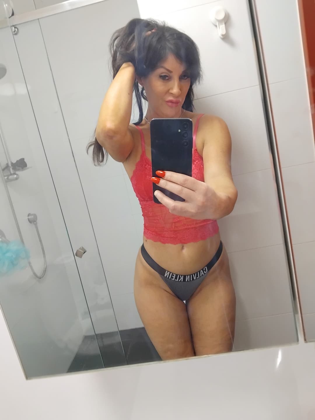 A woman takes a mirror selfie in a bathroom, wearing a red lace top and black Calvin Klein underwear. Evoking the allure of an erotic massage, she holds her phone up with one hand and touches her hair with the other.