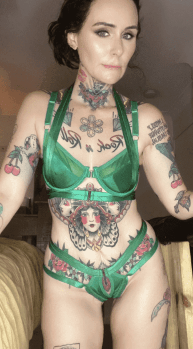 A woman with dark curly hair and prominent colourful tattoos on her arms, chest, and torso poses indoors beside a bed in a warmly lit room, evoking the sensual ambience of a milking table massage, while wearing a green lingerie set.