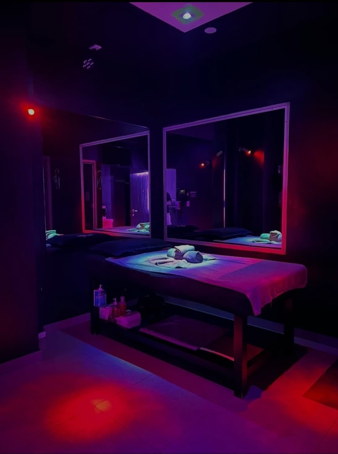 Soft purple and red lighting bathes a massage room at Mistiques Newtown, highlighting the towel-covered table and mirrored walls.