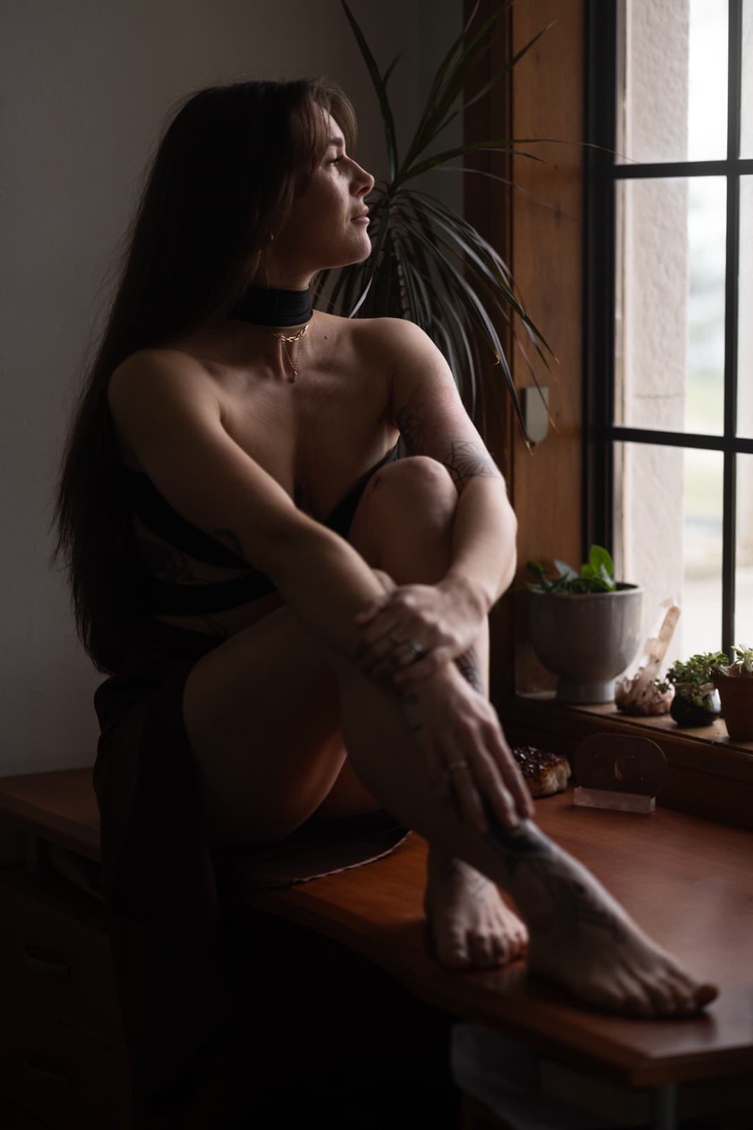 A woman with long hair sits on a wooden desk by a window, looking outside. An independent masseuse in a dark dress and choker, she wraps one arm around her knees as houseplants and decorative items line the windowsill.
