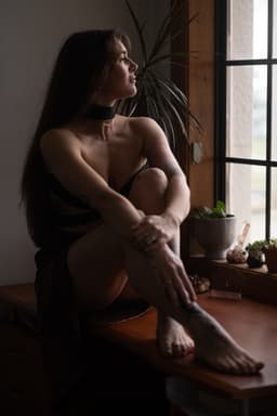 A woman with long hair sits on a wooden desk by a window, looking outside. An independent masseuse in a dark dress and choker, she wraps one arm around her knees as houseplants and decorative items line the windowsill.