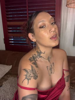 A person with short curly hair, facial piercings, and visible tattoos poses indoors on a brown sofa, wearing a red off-the-shoulder top. Red wooden shutters and a beige wall are in the background.