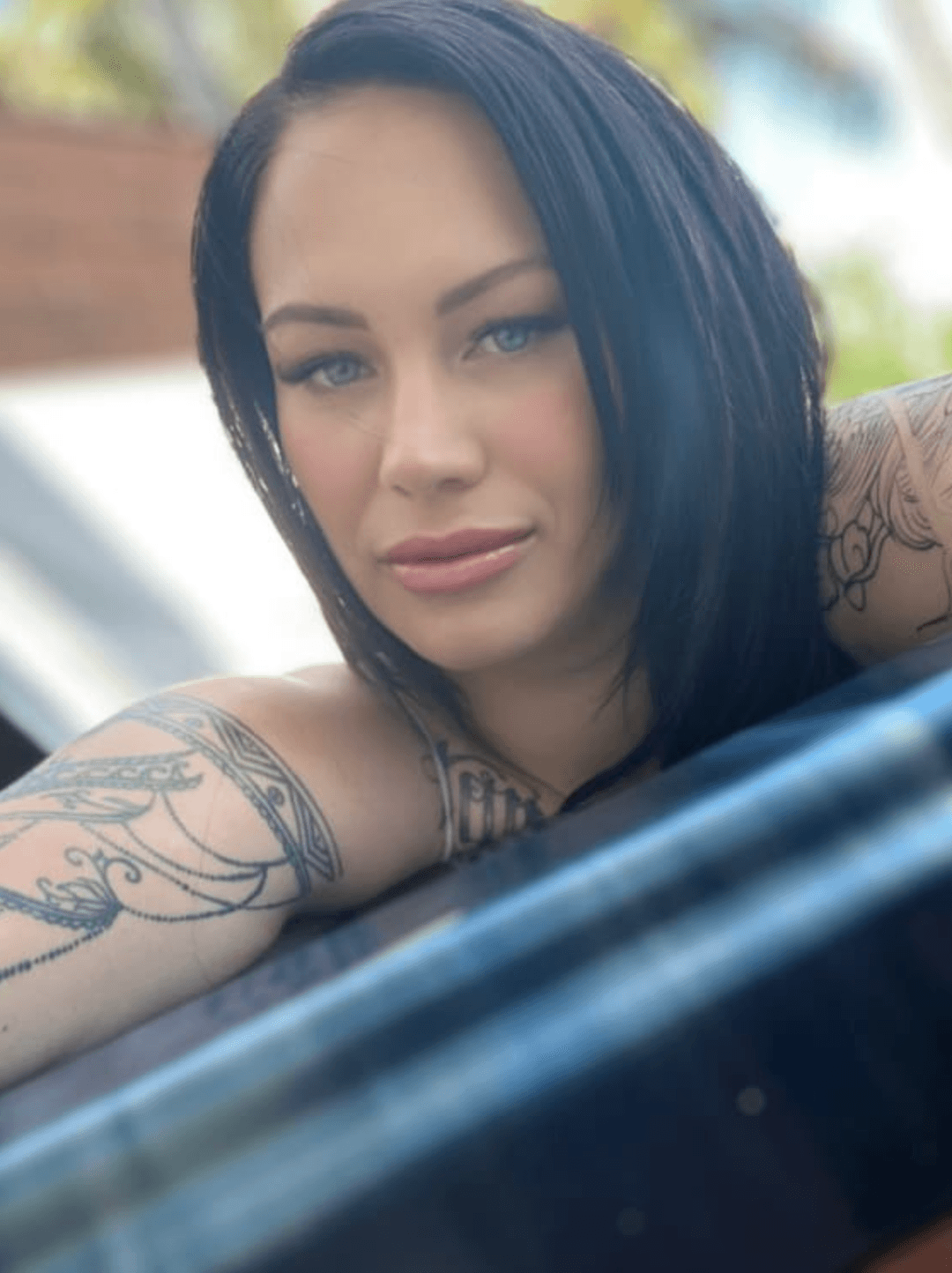 A woman with dark hair and tattoos on her arms looks into the camera and smiles slightly. The photo is taken outdoors in natural light, with a blurred background of greenery and buildings.