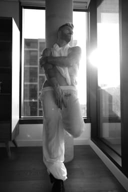 A person in sheer, loose clothing poses gracefully in front of a window with sunlight streaming in, one leg bent and arms folded over their chest, creating a dramatic, artistic scene in black and white.