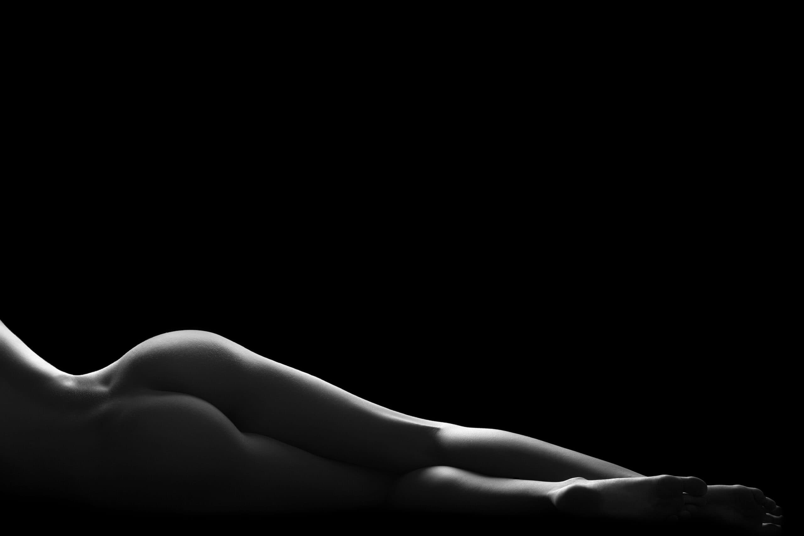 Black and white photo of a nude person lying on their side against a black background, their lower body and legs softly lit—capturing the artistic silhouette often celebrated in sensual massage imagery. Black and white photo of a nude person lying on their side against a black background, their lower body and legs softly lit—capturing the artistic silhouette often celebrated in sensual massage imagery.