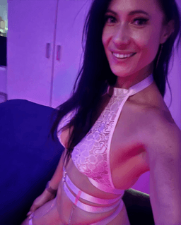 Tamara Rose smiling indoors under soft purple lighting while wearing a pink strappy top, expressing confident sensuality associated with tantric massage and embodied touch therapy services Tamara Rose smiling indoors under soft purple lighting while wearing a pink strappy top, expressing confident sensuality associated with tantric massage and embodied touch therapy services
